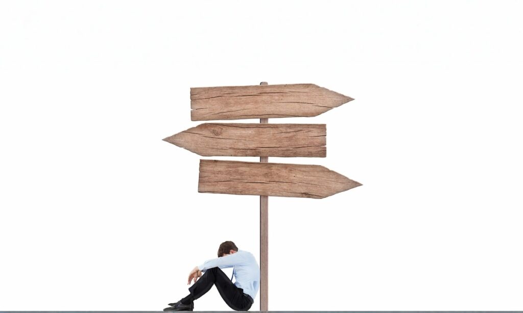 مردی خسته و بی‌انرژی که در پایین یک تابلوی چوبی چندجهته نشسته؛ نمادی از خستگی تصمیم‌گیری و فرسودگی ذهنی از انتخاب‌های متعدد.