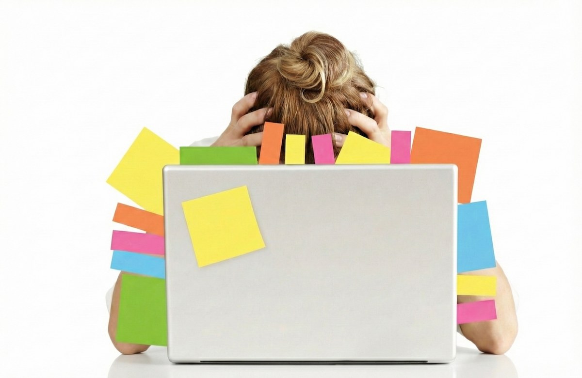 فریلنسر کلافه که سرش را میان دستانش گرفته و پشت لپ‌تاپ پوشیده از یادداشت‌های کاری پنهان شده؛ نمادی از فشار روانی چندگانه و تفاوت جنس فرسودگی شغلی در فریلنسرها نسبت به کارمندان.