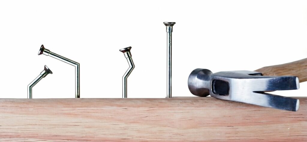 میخ‌های کج‌شده در اثر ضربه و چکشی که کنار یک میخ سالم رها شده است؛ تصویر مفهومی برای درماندگی آموخته‌شده و توقف تلاش پس از شکست.